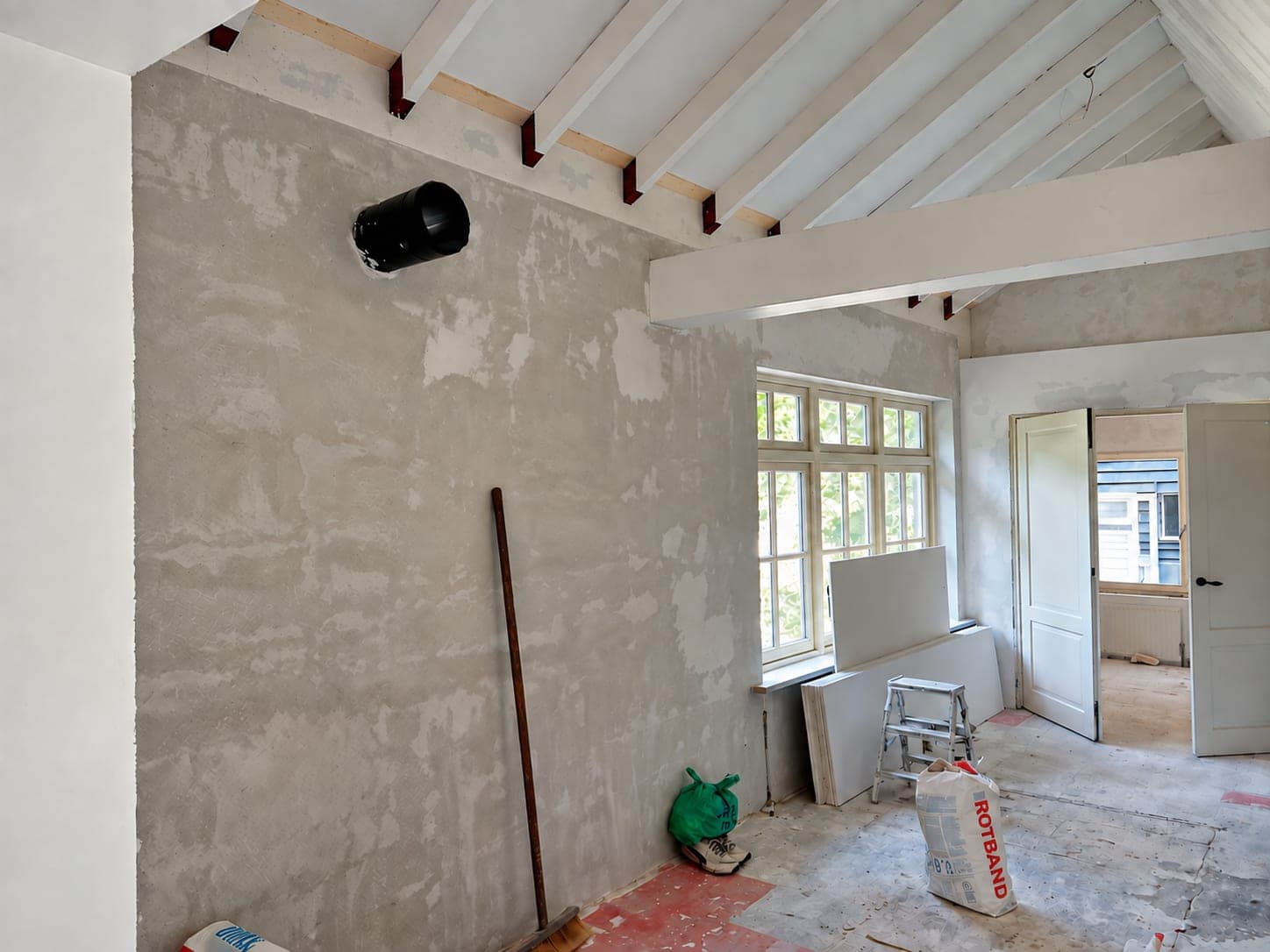 Voor: kamer onder de dakbalken met een kale, deels gerepareerde wand en bouwmateriaal op de vloer